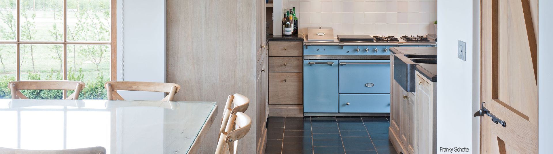 Helle Küche im Landhausstil mit Holzschränken, Esstisch mit Glasplatte, großen Fenstern und blauem Retro-Standherd
