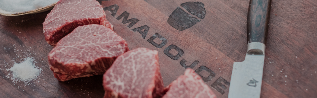 Mehrere rohe Rindersteaks liegen auf einem Holzschneidebrett von Kamado Joe, daneben ein Messer und etwas Salz