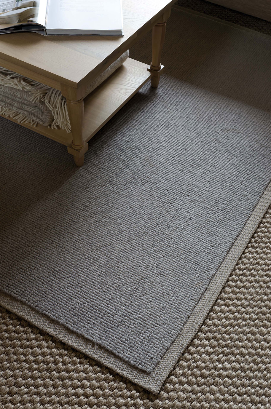 Grau gestrickter Teppich aus Naturfasern liegt unter einem hellen Holztisch auf einem weiteren strukturierten Teppich