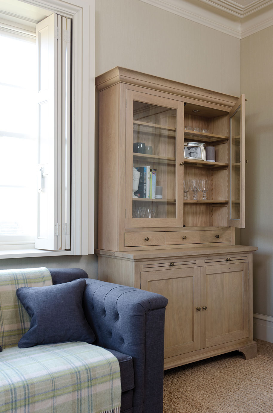 Vitrinenschrank aus hellem Holz mit Glastüren, teilweise geöffnet, gefüllt mit Gläsern, Büchern und Dekoration, daneben ein dunkelblaues Sofa mit Kissen.