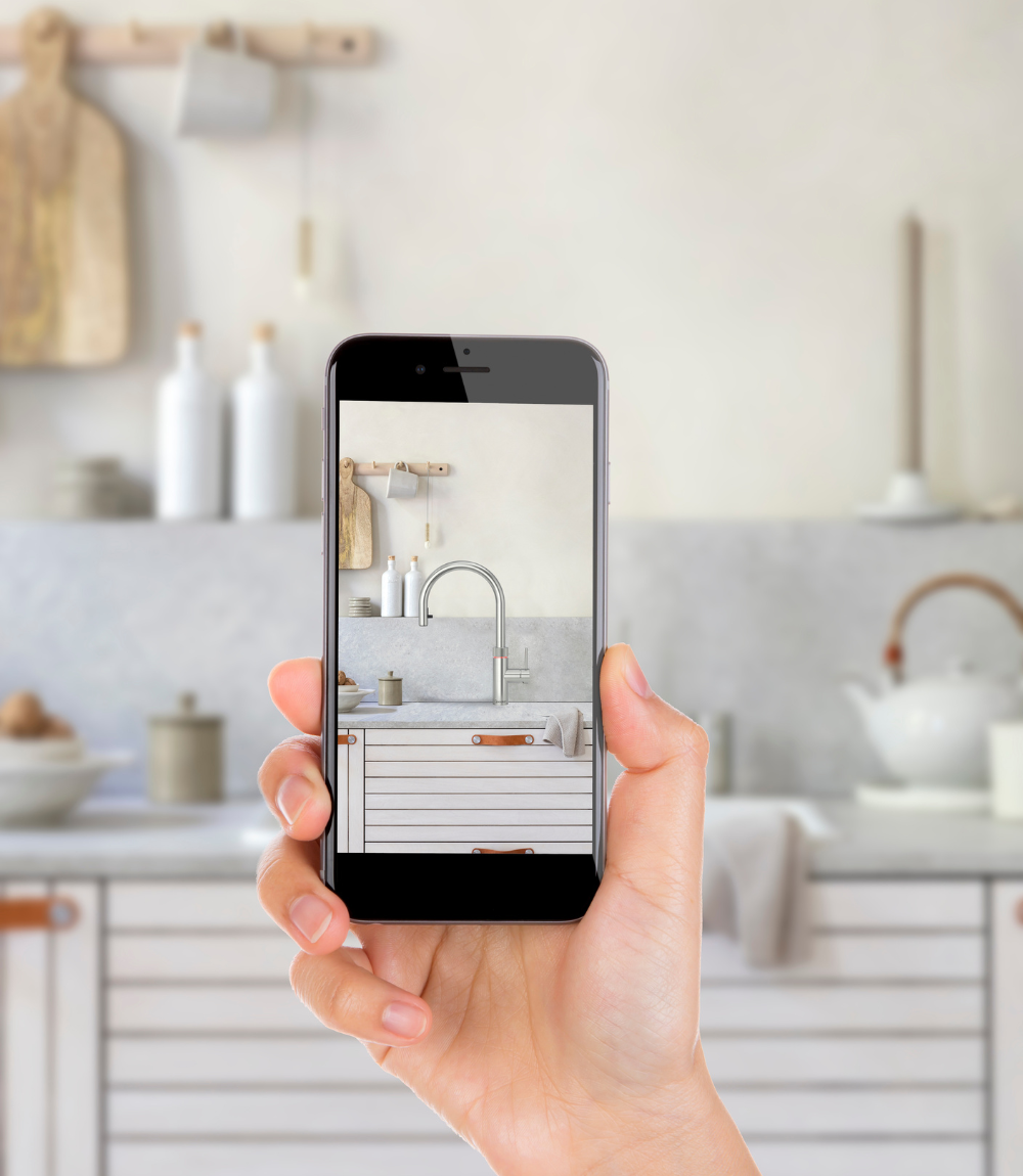 Nahaufnahme einer Hand, die ein Smartphone hält, auf dessen Display eine moderne, minimalistische Küche mit Wasserhahn und hellen Holzelementen zu sehen ist
