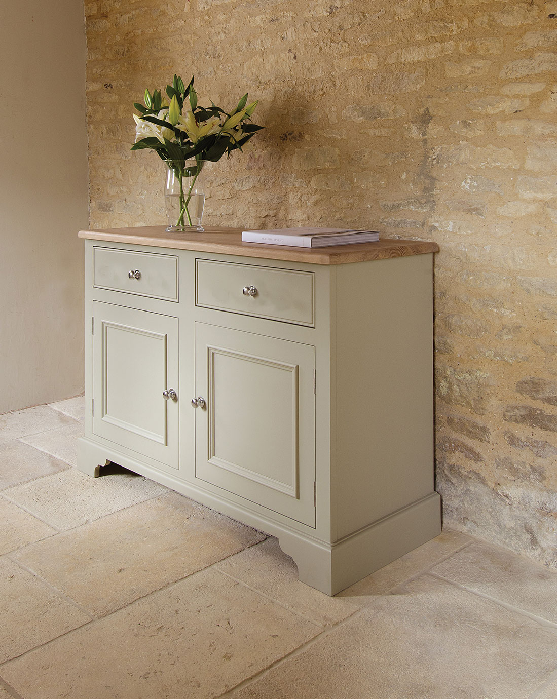 Cremfarbenes Sideboard im Landhausstil mit Holzplatte, zwei Schubladen und zwei Türen, darauf eine Glasvase mit weißen Lilien und ein Buch, vor einer Natursteinwand stehend
