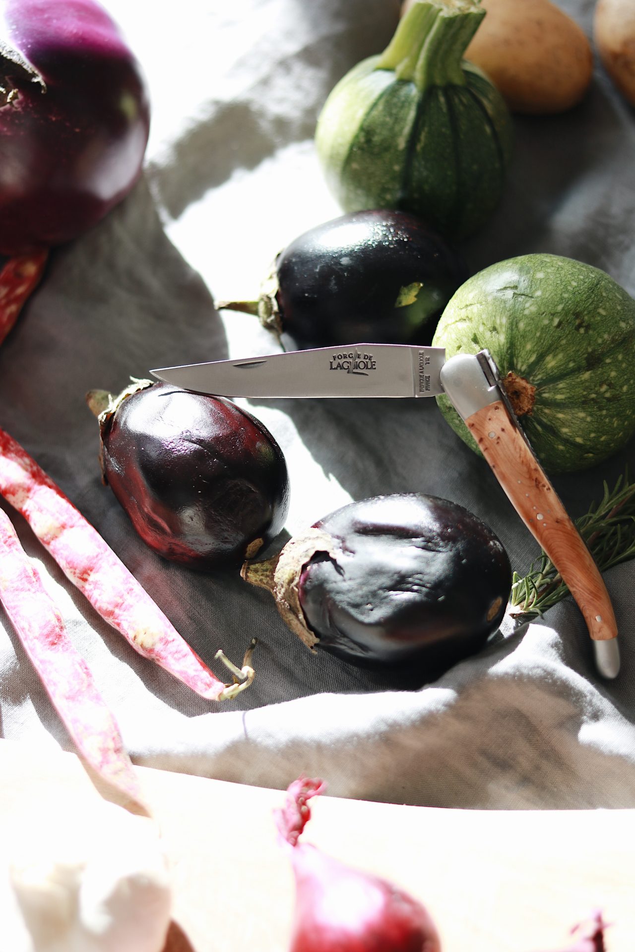 Verschiedene Gemüsesorten, darunter Auberginen, runde Zucchini, Kartoffeln, Bohnen und ein Kräutertzweig, neben einem hochwertigen Klappmesser auf einem hellen Tuch