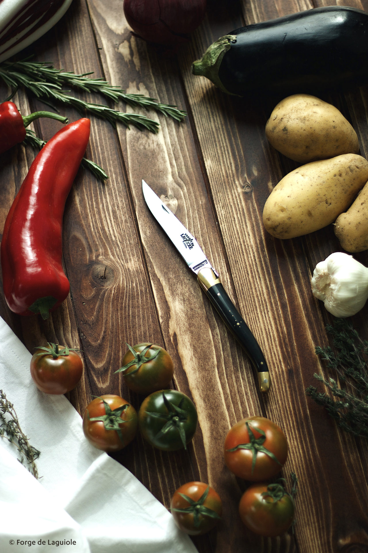 Elegantes Küchenmesser von Forge de Laguiole umgeben von frischem Gemüse wie Tomaten, Kartoffeln, Paprika, Aubergine, Kräutern und Knoblauch auf einem Holzuntergrund