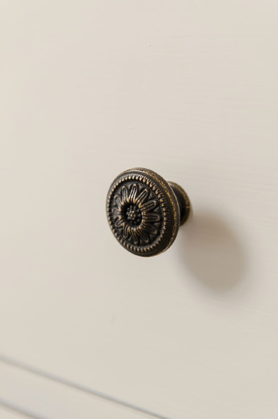 Detailaufnahme eines antik wirkenden, runden Möbelknaufs aus Metall mit floralem Muster an einer hellen Schublade