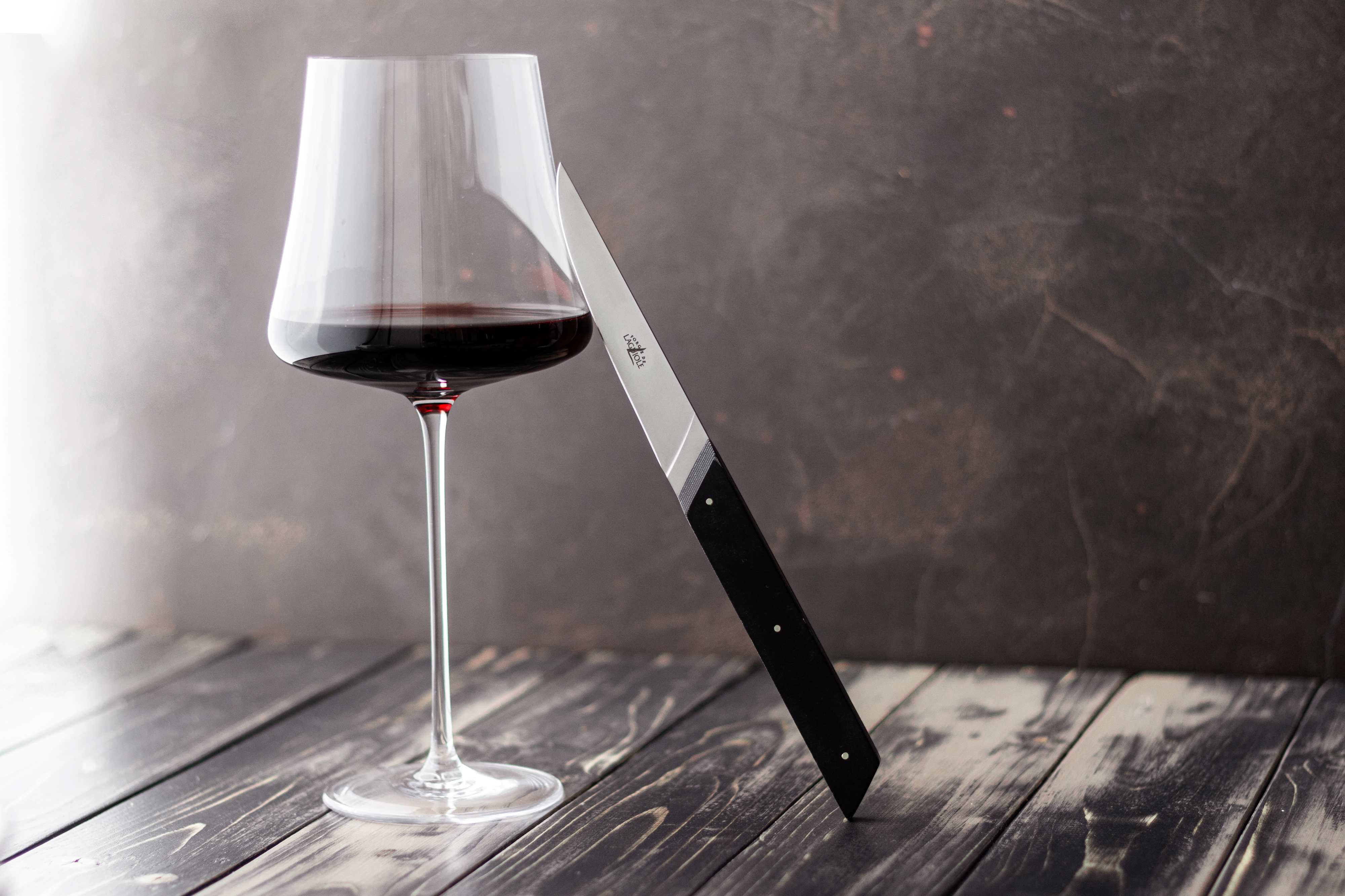 Ein halb gefülltes Weinglas mit Rotwein und ein Messer mit schwarzem Griff, das am Glas lehnt, auf einem dunklen Holztisch