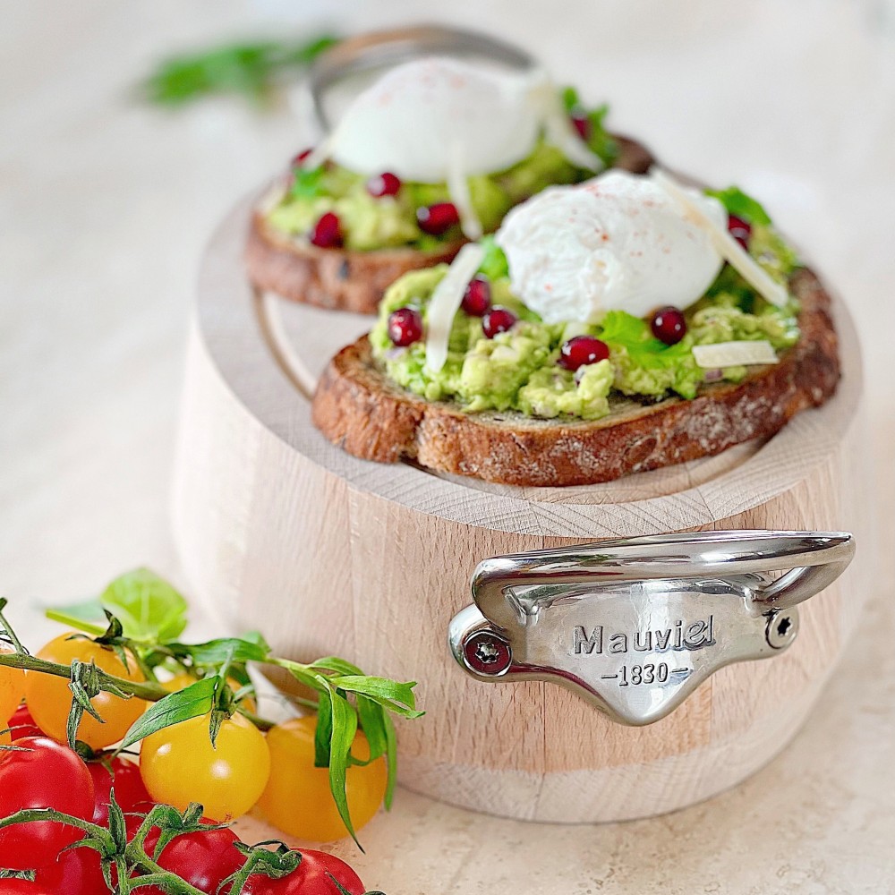 Zwei Avocado-Toasts mit pochiertem Ei, Granatapfelkernen und Käse auf einem Holzbrett, daneben frische gelbe und rote Tomaten