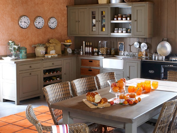 Einladende Landhausküche mit Holzmöbeln, offenen Regalen, mehreren Uhren an der Wand und einem großen Esstisch, auf dem Frühstück mit Orangensaft und Gebäck serviert wird.