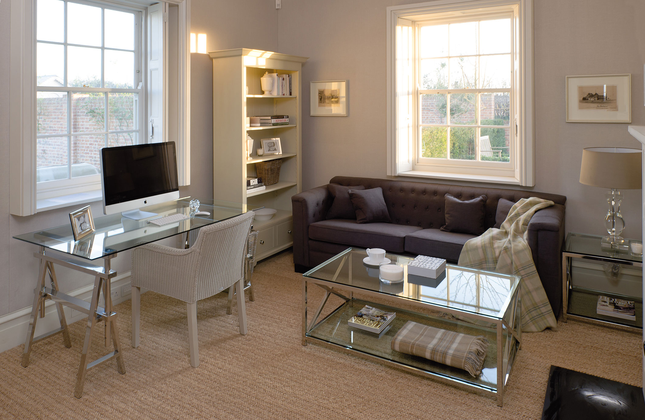 Helles Wohnzimmer mit Glas-Schreibtisch, Computer, Sessel, dunkelgrauem Sofa, Couchtisch und Bücherregal in elegantem Design