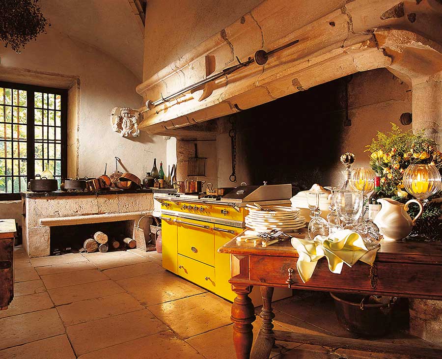 Gemütliche Landhausküche mit großem gelben Herd, Steinkamin, Fenstergitter und gedecktem Holztisch mit Geschirr und Blumen