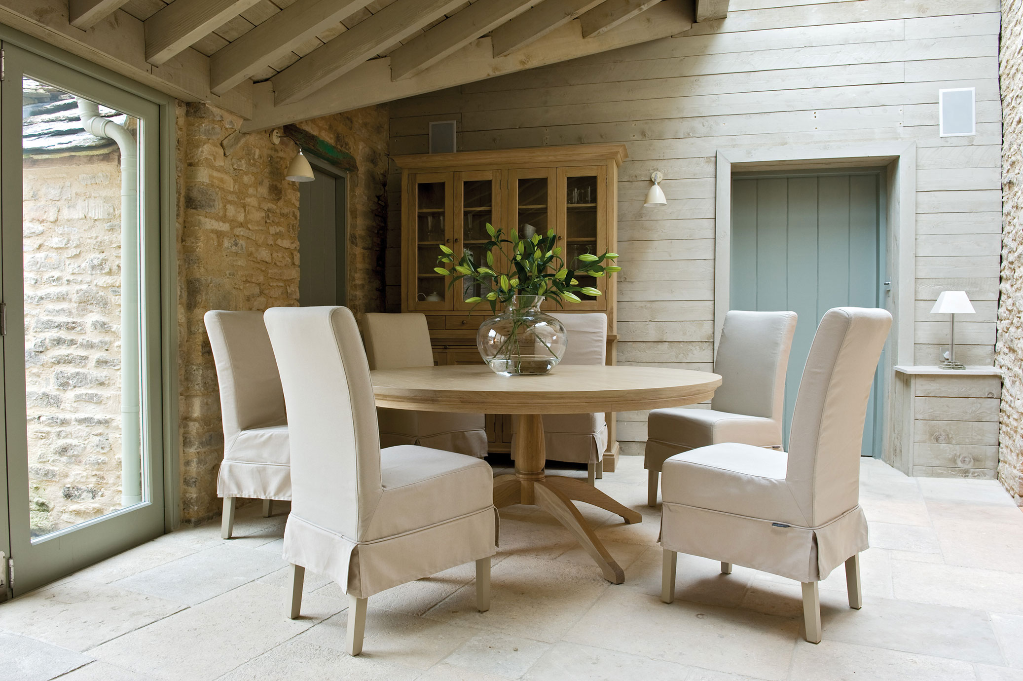 Helles Esszimmer mit rundem Holztisch, sechs beige Stühlen und Vase mit grünen Zweigen, gemütlich im Landhausstil eingerichtet