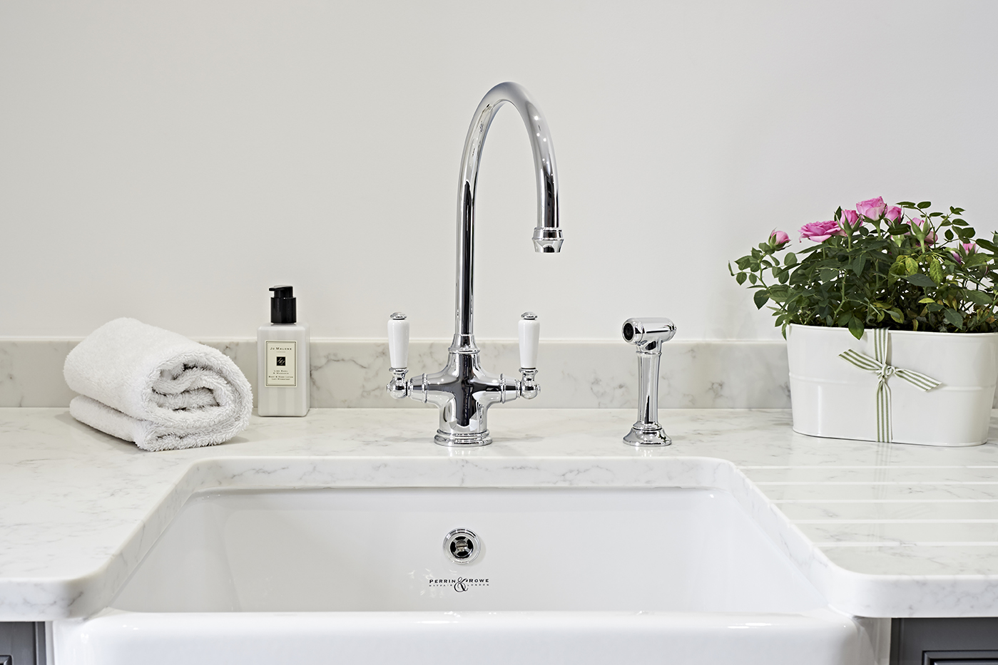 Waschbecken aus Keramik mit glänzendem Chrom-Wasserhahn, weißem Handtuch, Seifenspender und Blumentopf mit rosa Blumen auf einer Marmorarbeitsplatte