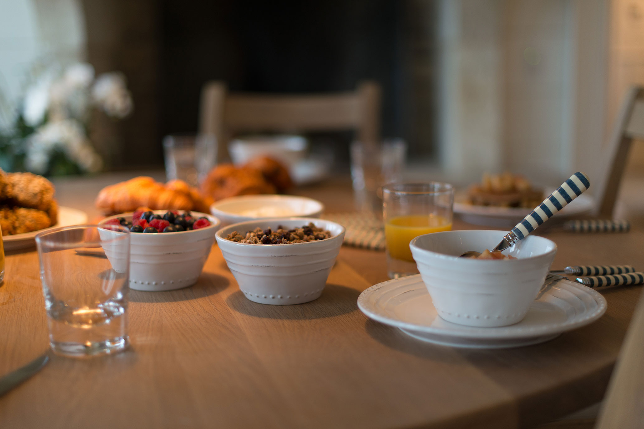 Ein gedeckter Frühstückstisch mit weißen Schalen, gefüllt mit Müsli, Beeren und Joghurt, daneben Gebäck und einem Glas Orangensaft in gemütlicher Atmosphäre