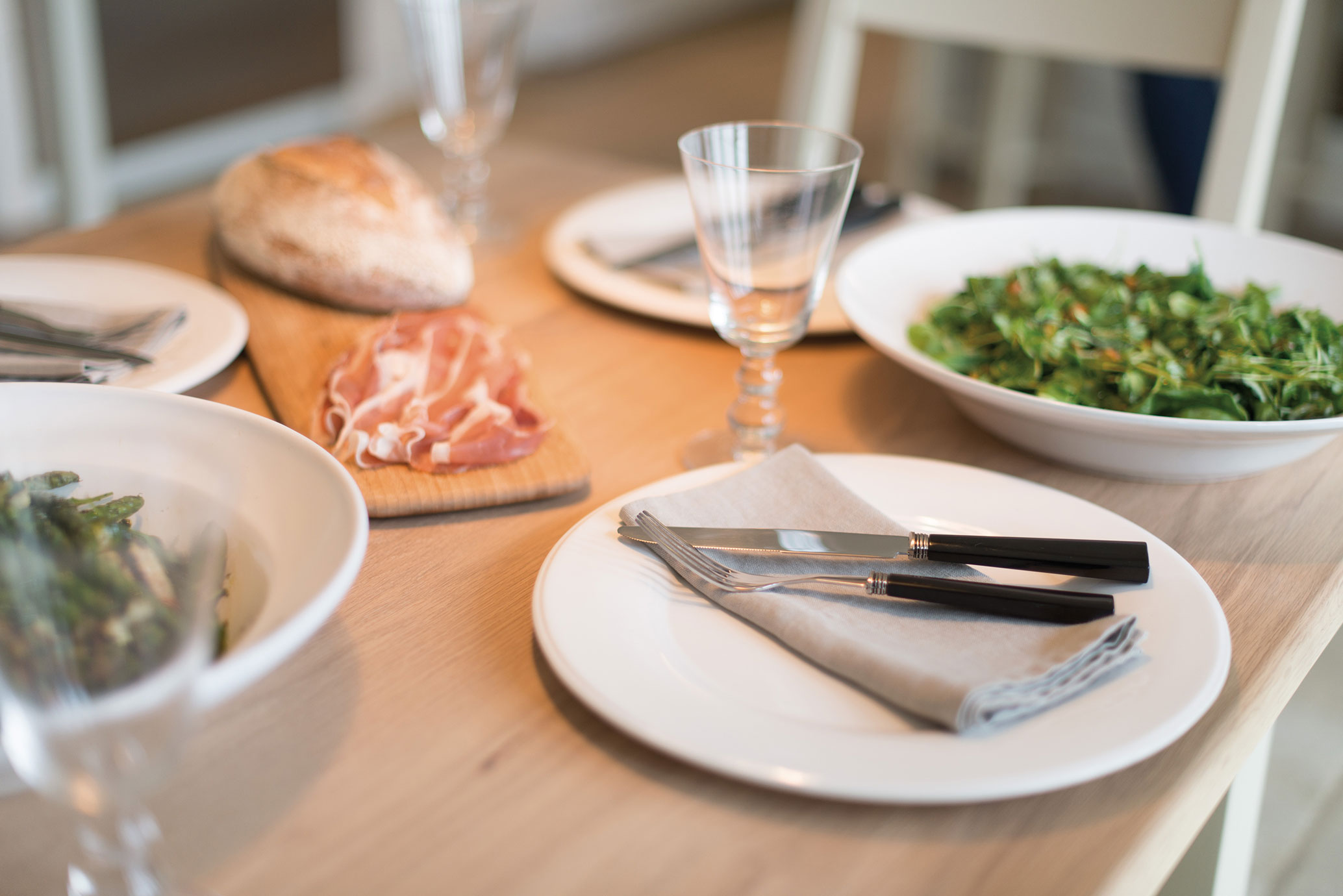 Nahaufnahme eines gedeckten Esstisches mit weißem Geschirr, Besteck, Glas, einer Schale Salat, einem Holzbrett mit Brot und Aufschnitt.