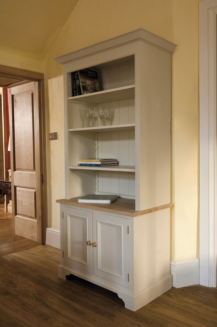 Cremfarbener Buffetschrank im Landhausstil mit offenen Regalen, Gläsern und Büchern, darunter Schranktüren und Holzplatte in einem gemütlichen Raum