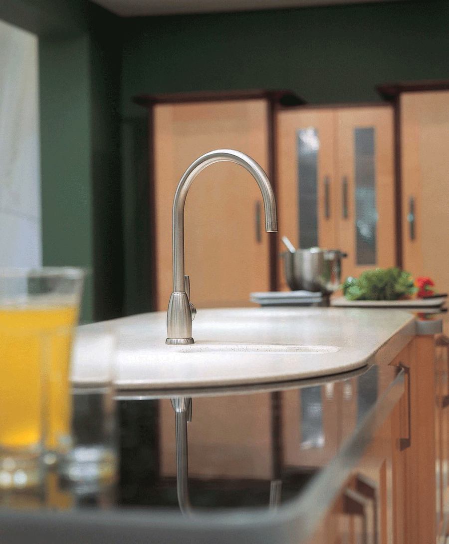 Ein moderner Edelstahl-Küchenwasserhahn installiert auf einer hellen Kücheninsel, im Hintergrund Holzschränke, ein Glas Saft und frisches Gemüse auf der Arbeitsfläche.