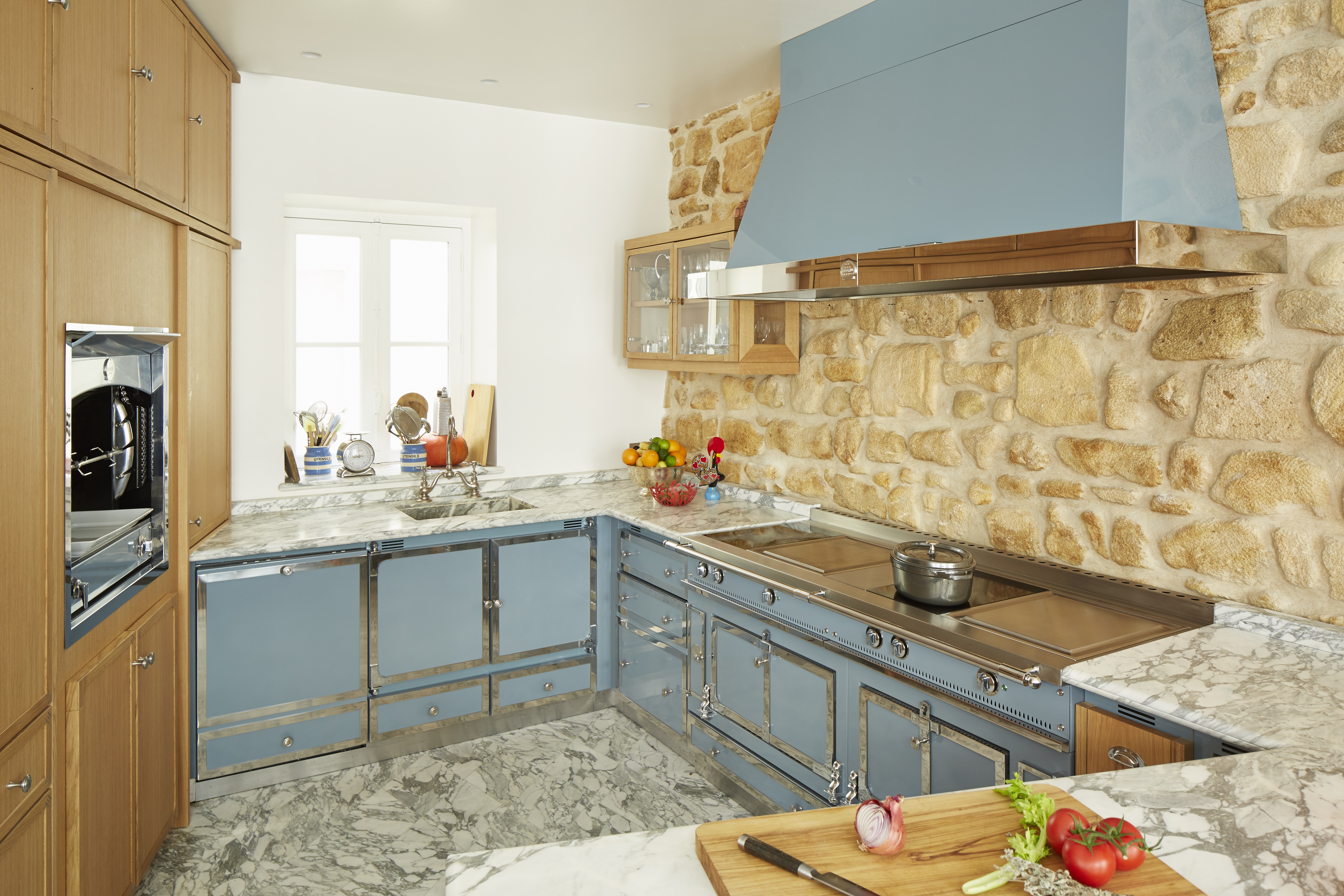 Helle Küche mit blauen Unterschränken, Marmorarbeitsfläche, Steinwand, Einbauofen und frischem Gemüse auf dem Schneidebrett