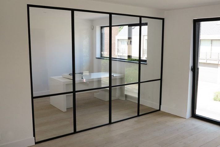 Raum mit moderner Glaswand und schwarzen Metallrahmen als stilvoller Raumteiler, dahinter ein Schreibtisch vor großem Fenster