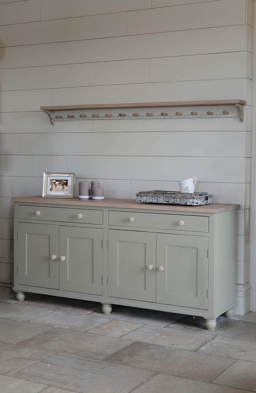 Schlichtes graues Sideboard aus Holz vor einer weißen Wand mit Ablage und Hakenleiste, dekoriert mit Bilderrahmen, Kerzen und geflochtenem Korb