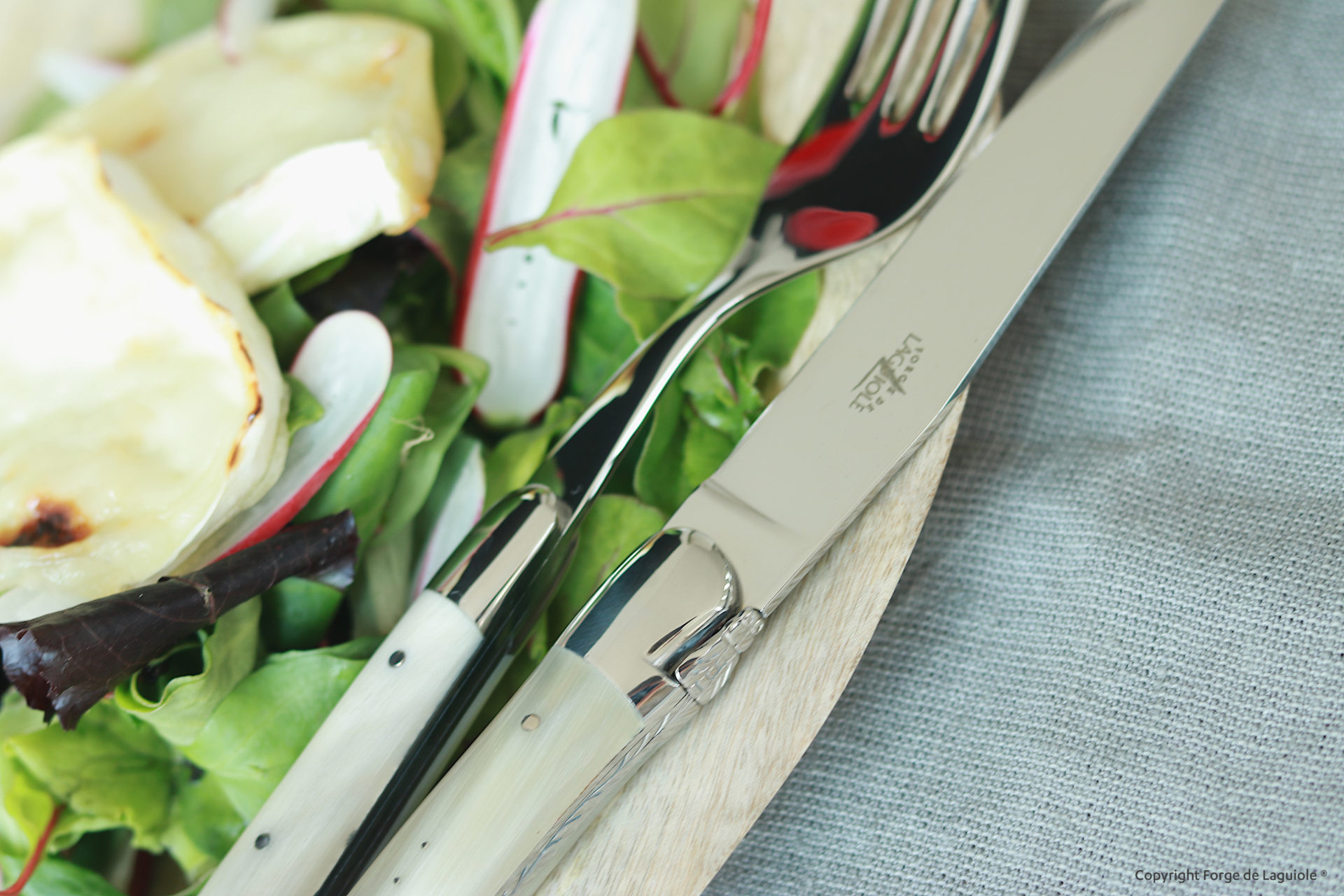 Laguiole Messer und Gabel mit hellem Griff, platziert auf einem Teller mit grünem Salat, Radieschen und gebackenem Ziegenkäse
