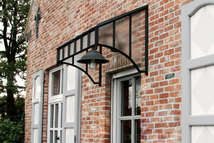 Modernes Glas- und Metallvordach mit schwarzem Rahmen und integrierter Außenlampe, montiert über einer Haustür an einer Fassade aus roten Ziegelsteinen