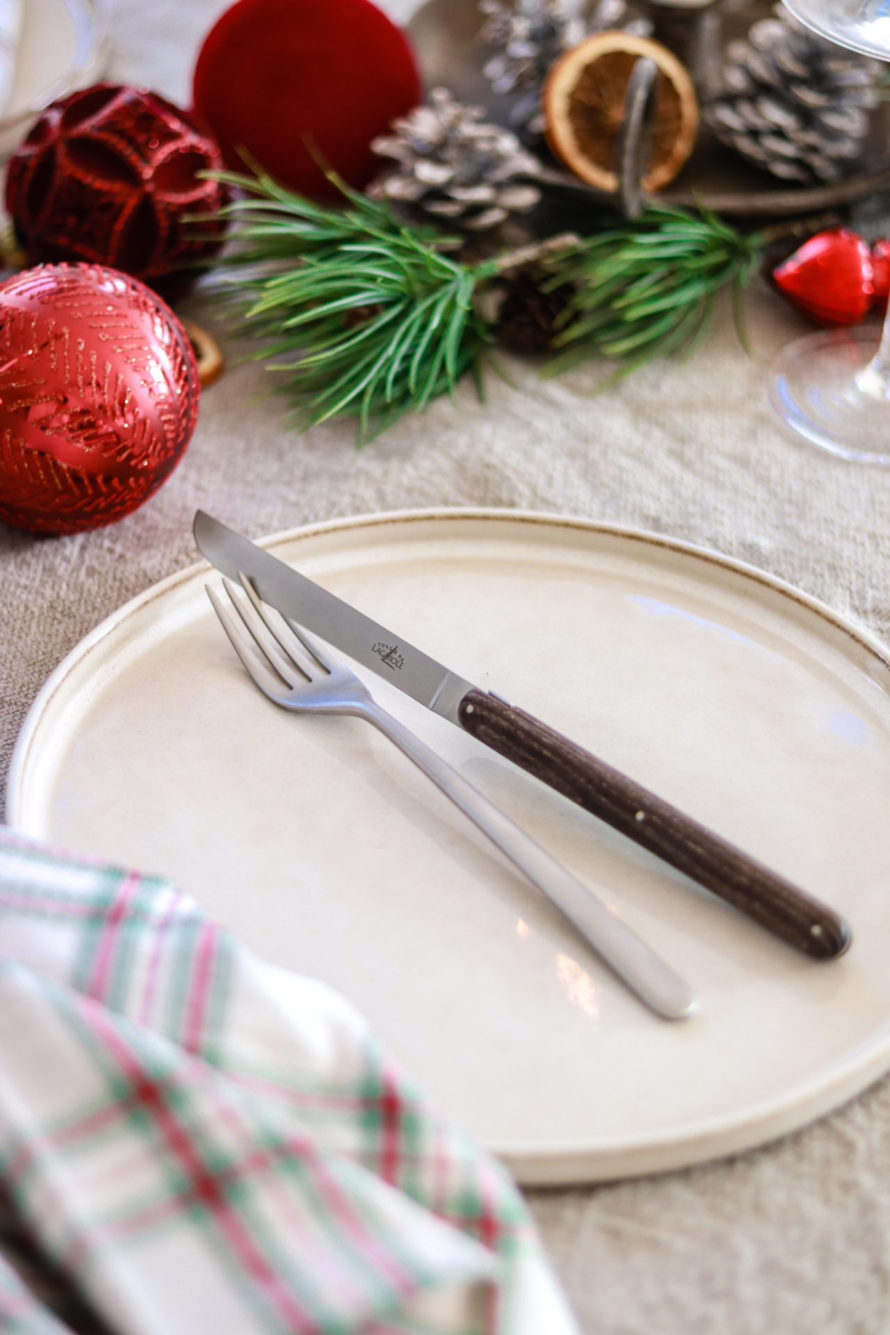 Weißer Teller mit Messer und Gabel, festlich gedeckt mit roter Weihnachtsdeko, Kieferzweigen und Zapfen