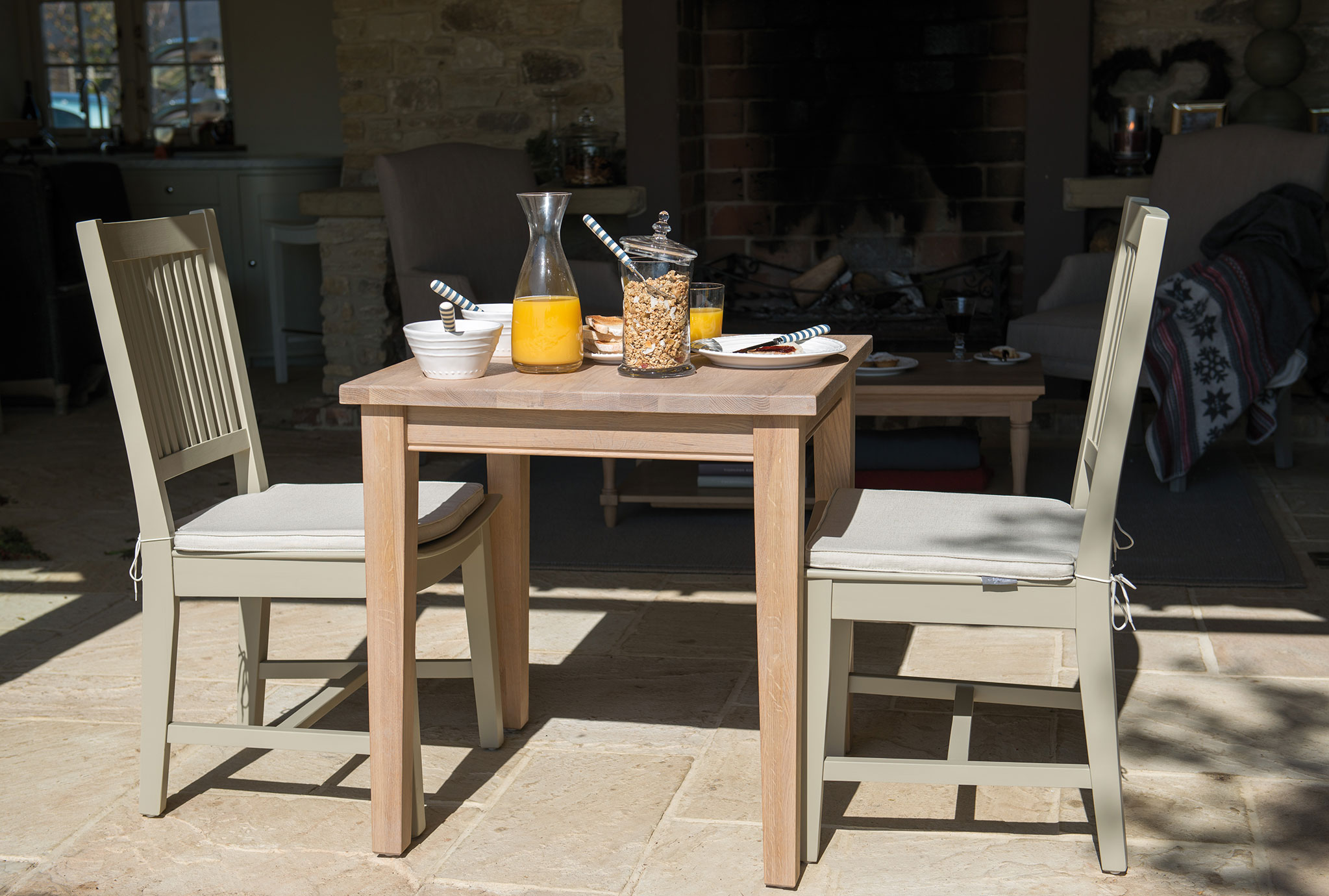 Holztisch mit zwei gepolsterten Stühlen, darauf Karaffe mit Orangensaft, Müslischale, Frühstücksgeschirr und Granola im Glas, sonnige Atmosphäre