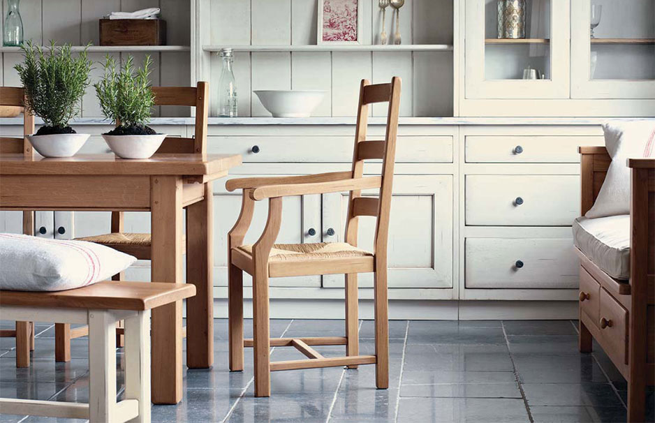 Gemütliches Esszimmer mit Holztisch, Holzstühlen, Bank, weißen Kissen und Küchenzeile im Landhausstil, verziert mit Schalen und Pflanzen auf dem Tisch.