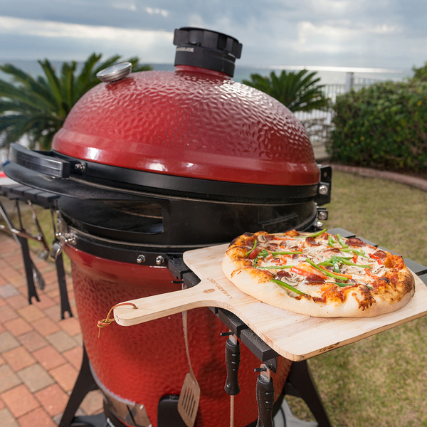 Pizza mit Gemüsebelag auf Holzschaufel, präsentiert vor einem roten Keramikgrill im Freien