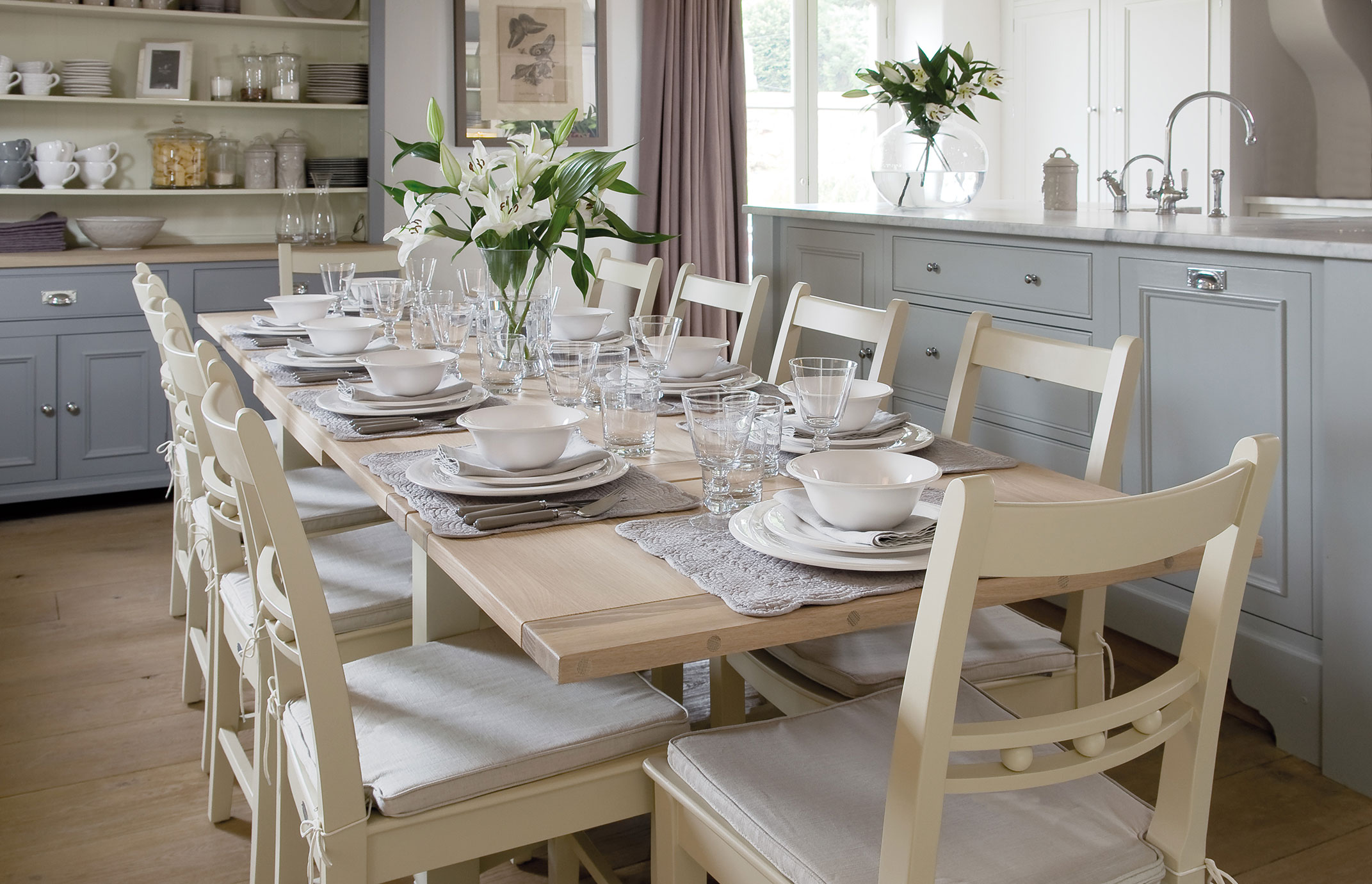 Heller Esstisch für acht Personen, eingedeckt mit weißem Geschirr, Gläsern, Stoffservietten und Blumenvase, in moderner Landhausküche