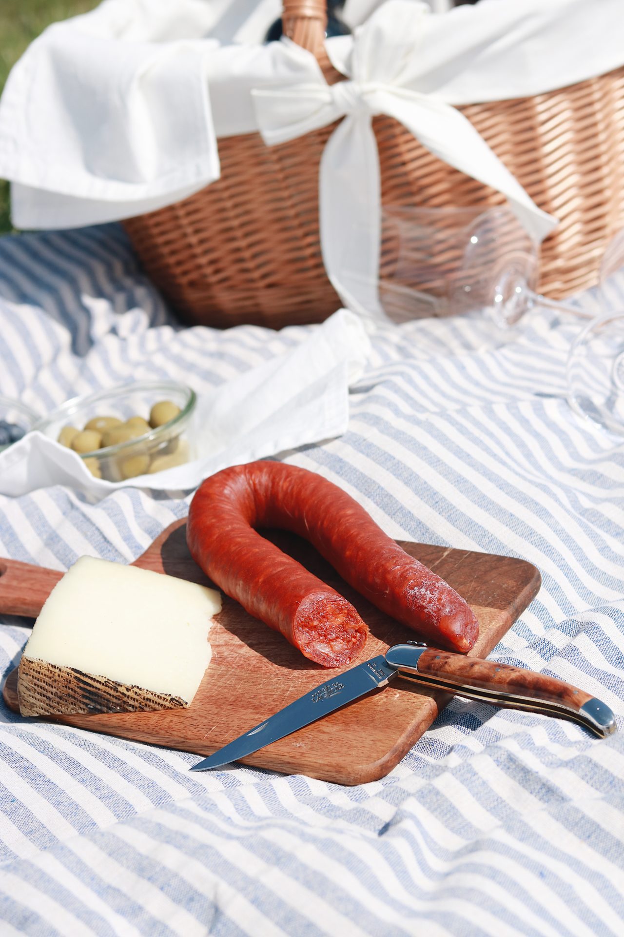 Picknick-Szene mit einem Holzbrett, darauf ein Stück Käse, eine Salami und ein Messer, daneben Oliven und ein geflochtener Picknickkorb auf einer gestreiften Decke