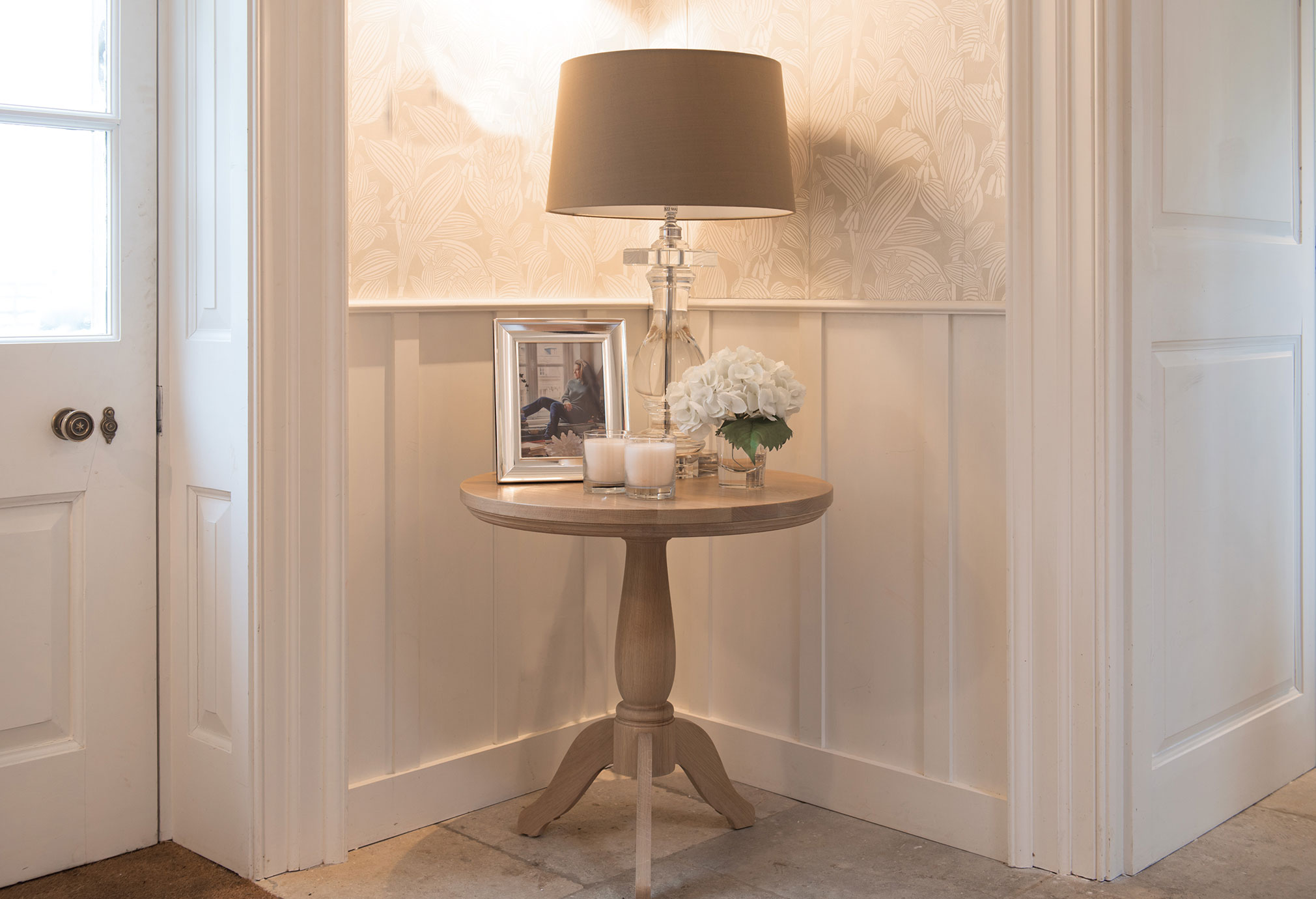 Runder Holztisch in einer Ecke mit Tischlampe, Bilderrahmen, weißen Blumen und Kerzen auf hellem Hintergrund
