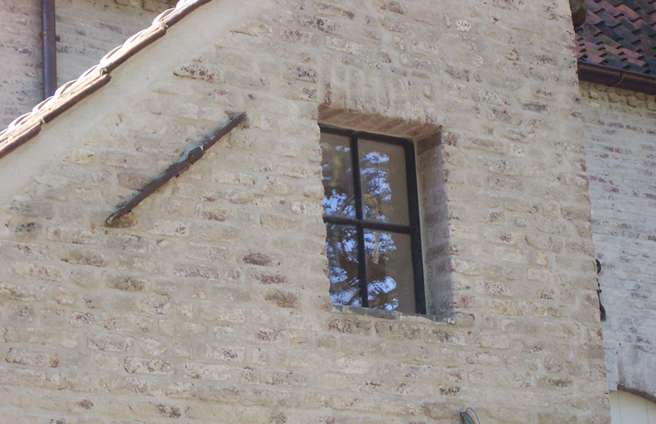 Quadratisches Fenster mit schwarzem Rahmen in einer alten Ziegelsteinwand, teilweise Spiegelung von Bäumen sichtbar