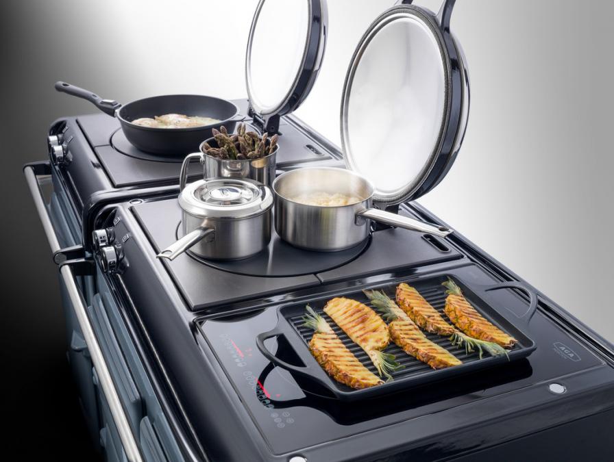 Ein moderner Herd mit geöffneten Kochplatten, verschiedenen Töpfen, Pfanne mit Gemüse und Grillpfanne mit Hähnchenfilets