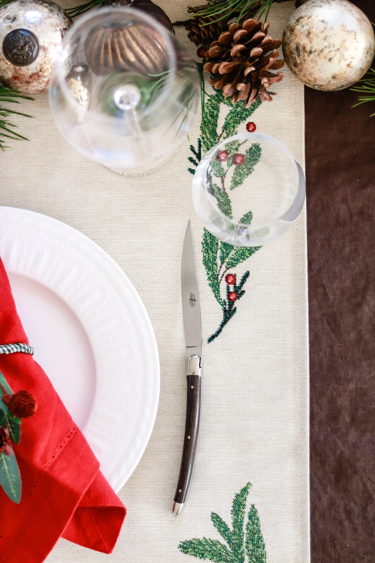 Weihnachtlich dekorierter Tisch mit Keramik-Teller, roten Servietten, Glas, Messer, Tannenzapfen und besticktem Tischläufer mit Zweigen