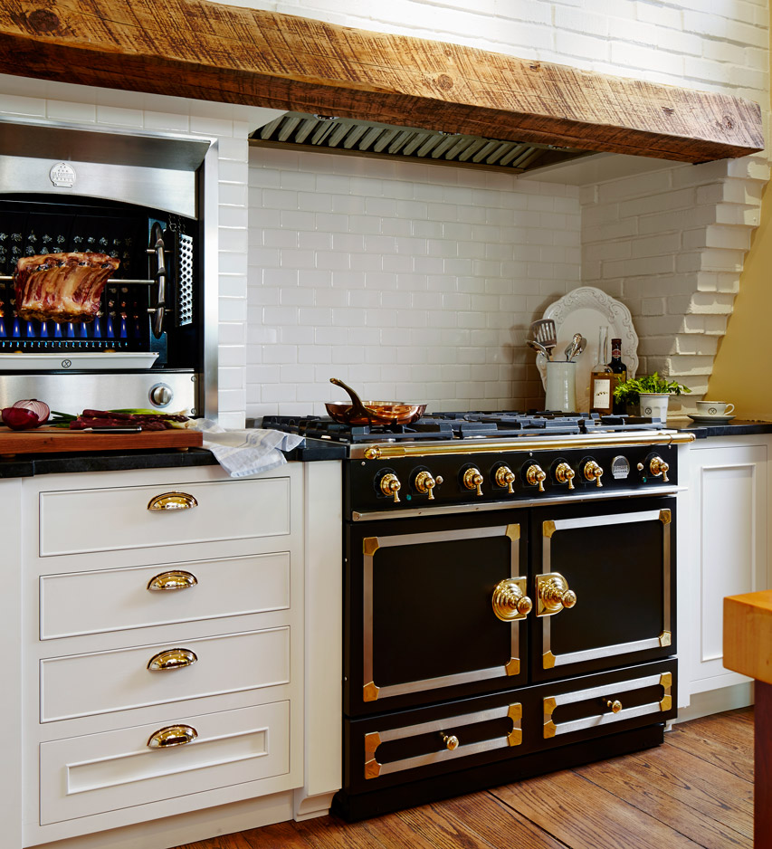 Elegante Küche mit schwarzem Herd und goldenen Bedienelementen, Grillstation mit Braten, weißen Schränken und rustikalem Holzbalken über dem Herd