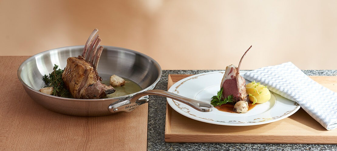 Lammkarree in Edelstahlpfanne mit Kräutern und Knoblauch, daneben ein angerichtetes Lammgericht mit Sauce und Kartoffelpüree auf einem Porzellanteller mit Stoffserviette