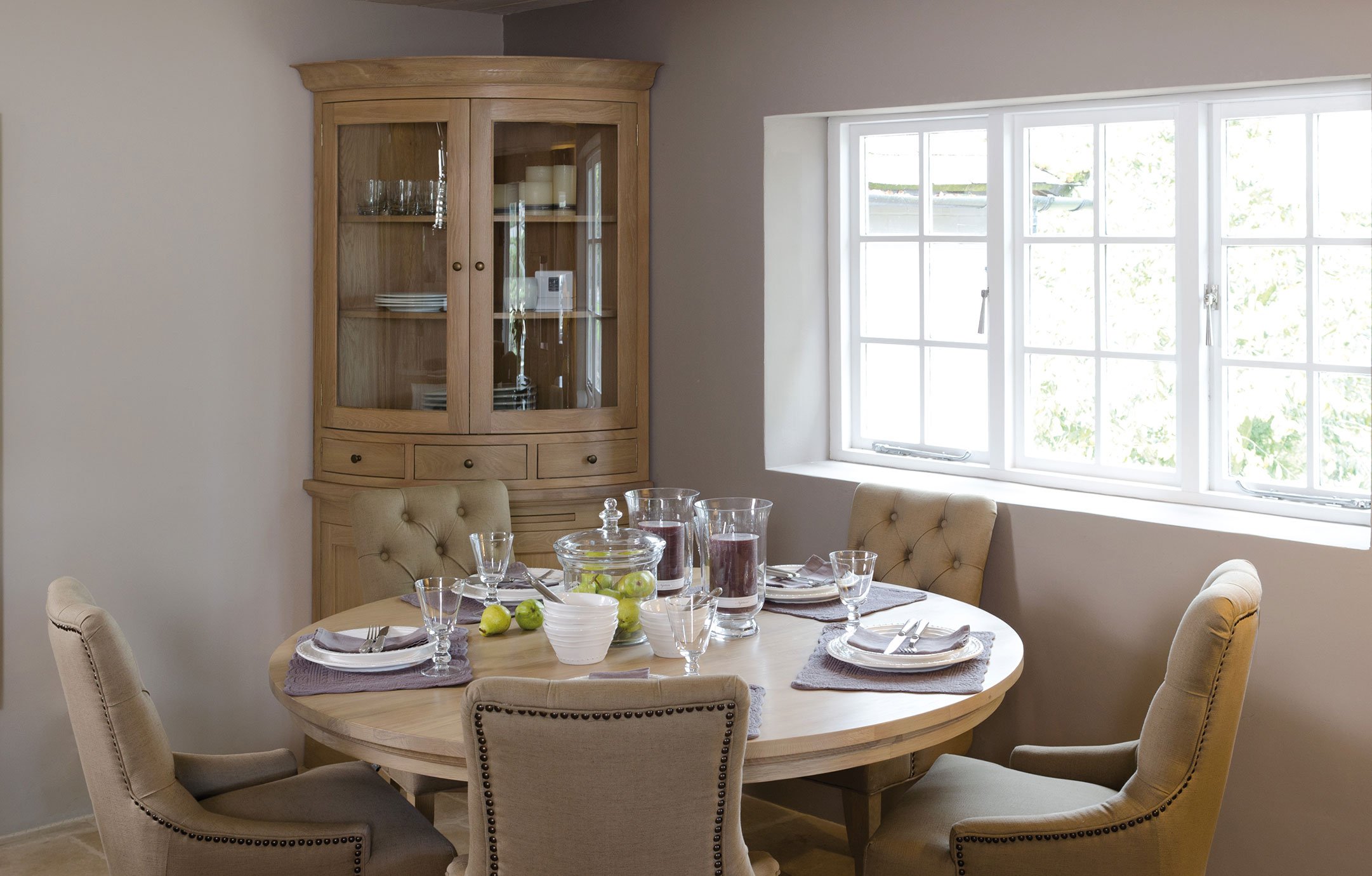 Modern eingerichtetes Esszimmer mit rundem Holztisch, beige gepolsterten Stühlen, dekoriertem Gedeck und einer Vitrine im Hintergrund