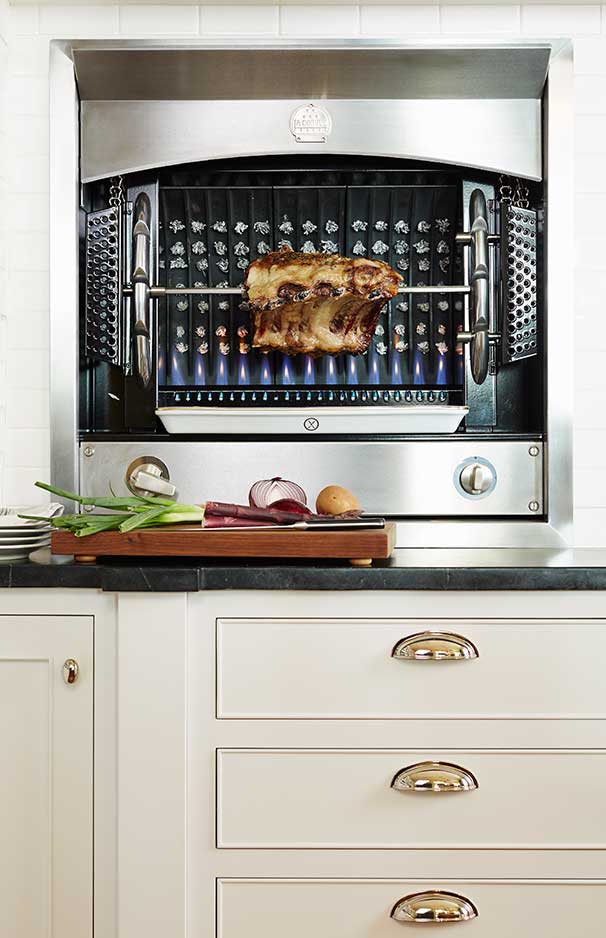Ein moderner Edelstahl-Gasgrill mit drehendem Spieß und saftigem Braten darüber, darunter frisches Gemüse auf einem Schneidebrett in einer eleganten Einbauküche.