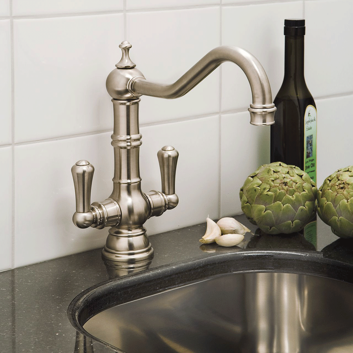 Küchenarmatur aus Edelstahl im klassischen Design neben Spüle, Artischocken, Knoblauch und Olivenölflasche auf schwarzer Arbeitsplatte