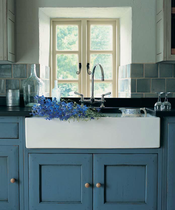 Weiße Keramikspüle im Landhausstil mit blauen Schränken, frischen blauen Blumen, Glasflaschen und Ausblick durch ein helles Fenster