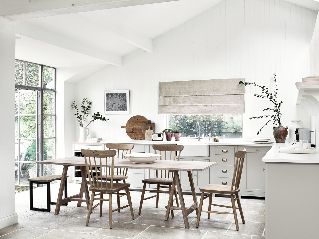 Helle, geräumige Landhausküche mit Holztisch, mehreren Holzstühlen, großen Fenstern, weißen Wänden, rundem Schneidebrett, Vasen mit Zweigen und modernem Küchenzubehör