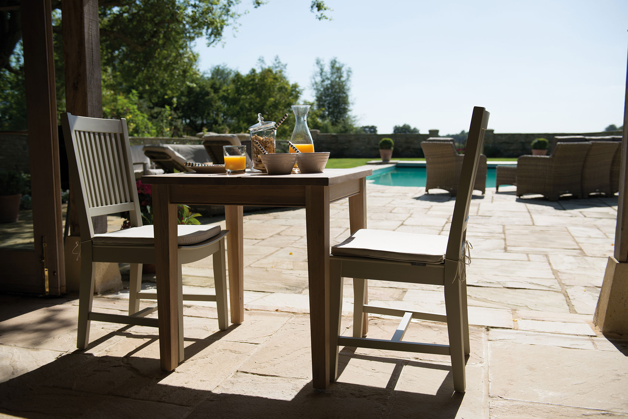 Holztisch mit zwei Stühlen, Frühstück mit Orangensaft und Müsli auf Terrasse neben Pool, Gartenmöbel und sonnige Atmosphäre