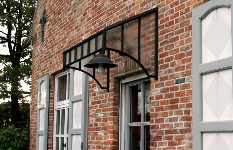Glas- und Metallvordach mit Außenlampe über einer grauen Haustür vor roter Backsteinwand