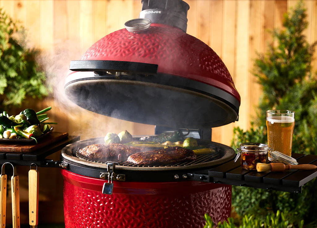 Roter Keramikgrill mit offenem Deckel, darauf Steaks und Gemüse, neben dem Grill stehen ein Glas Bier und Gewürze