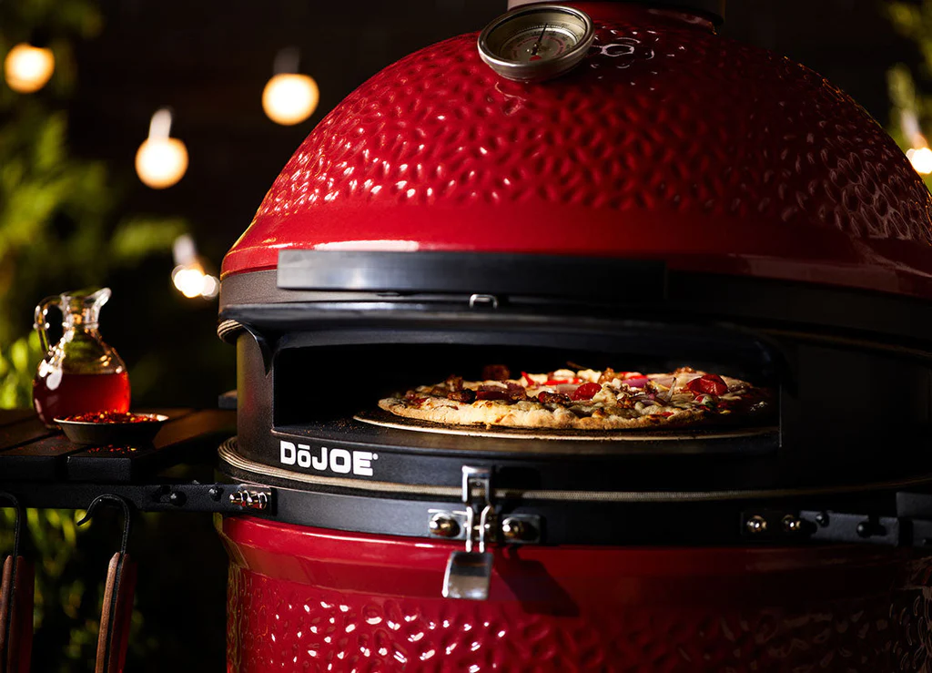 Roter Kamado Joe DoJoe Grill mit Pizza im Backvorgang, daneben eine Karaffe mit Öl und Gewürzen, stimmungsvolle Lichter im Hintergrund