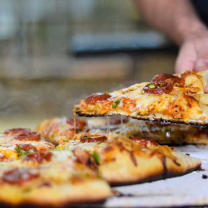 Hand nimmt ein Stück heiße Pizza mit geschmolzenem Käse, Salamischeiben und Gemüse vom ganzen Pizzafladen ab