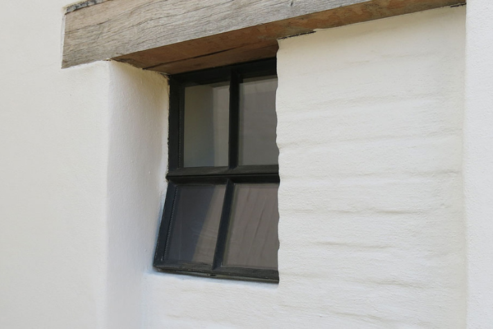 Schräg eingebautes, kleines Fenster mit schwarzem Rahmen in einer weiß verputzten Hauswand mit Holzbalken darüber