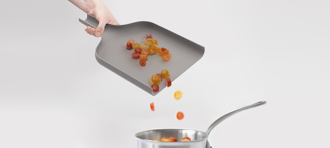Hand schüttet geschnittene Tomaten mithilfe einer grauen Schneidgut-Schaufel in einen Kochtopf aus Edelstahl