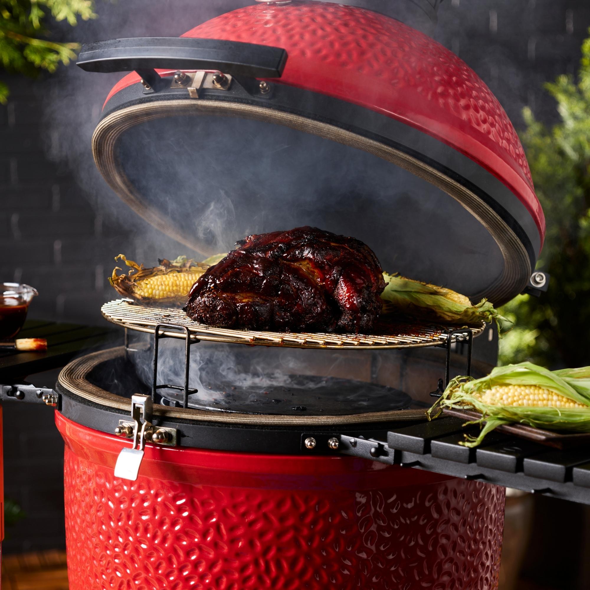 Ein roter Keramikgrill mit geöffnetem Deckel, auf dem ein großes Stück Fleisch und Maiskolben gegrillt werden, im Hintergrund leichte Rauchentwicklung.